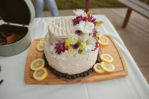 A rustic white frosted cake decorated with fresh daisies and purple flowers sits on a wooden board with lemon slices.
