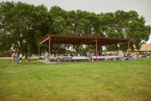 People gather under an open-air pavilion shelter with picnic tables on a grassy lawn surrounded by trees.