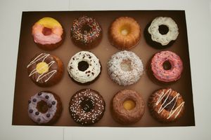 A dozen assorted gourmet donuts with various toppings including chocolate, vanilla, and pink frosting arranged on a brown background.