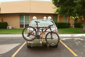 A bicycle mounted on a car's rear bike rack with a building and green trees in the background.