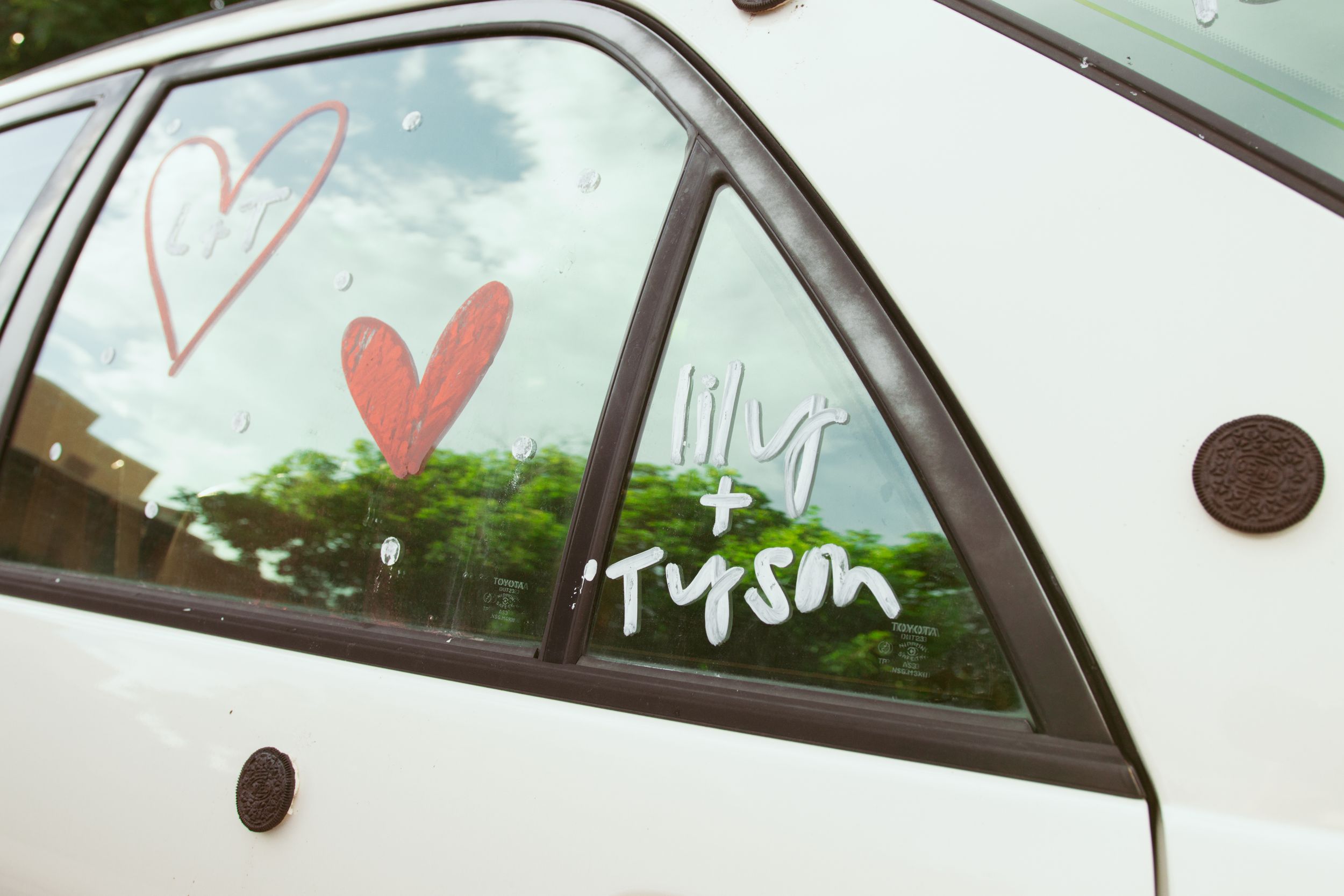 Hearts and writing drawn on a car window with white paint or marker against a green outdoor background.