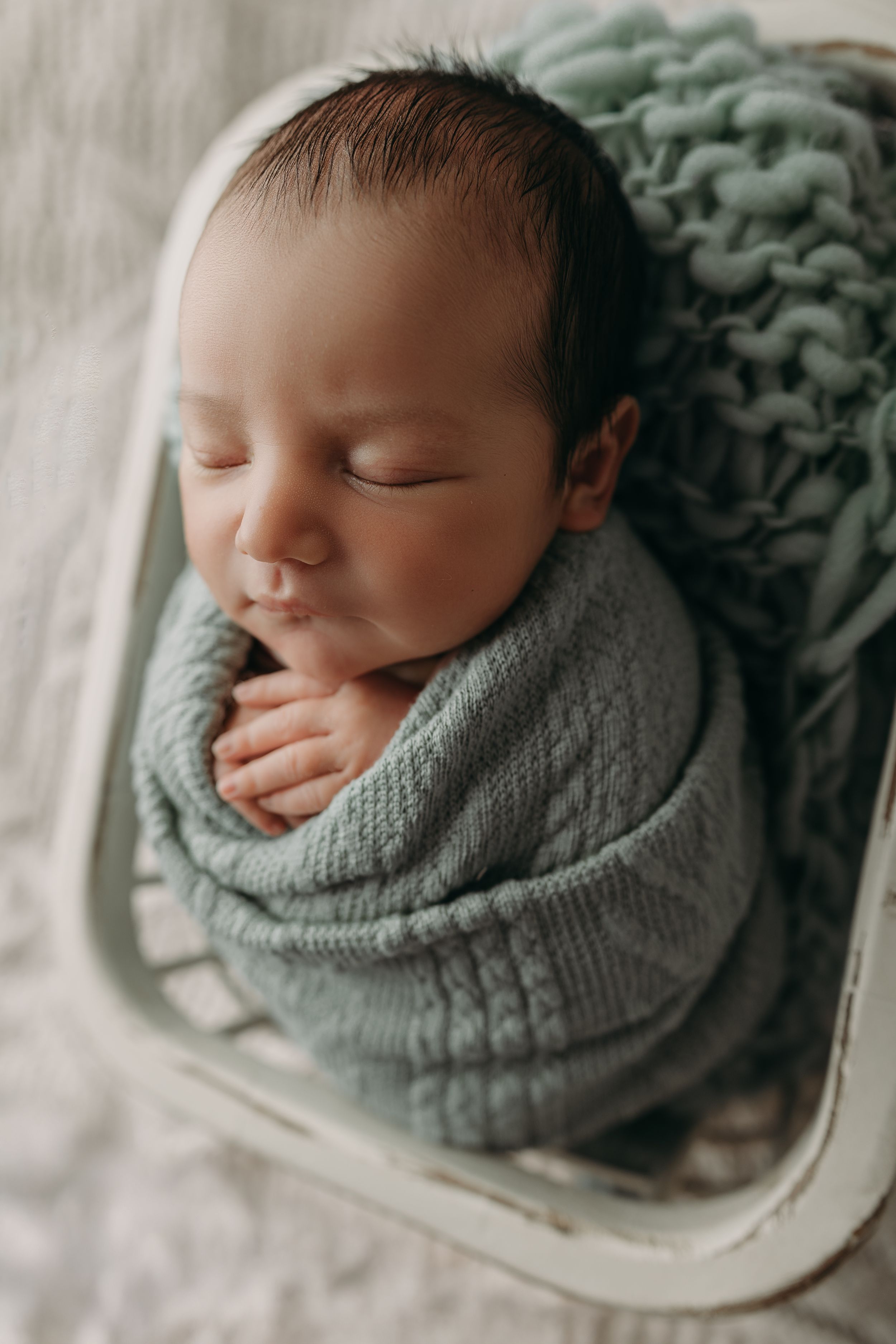 Peaceful sleeping newborn baby wrapped in soft sage green knit blanket laying in white basket.