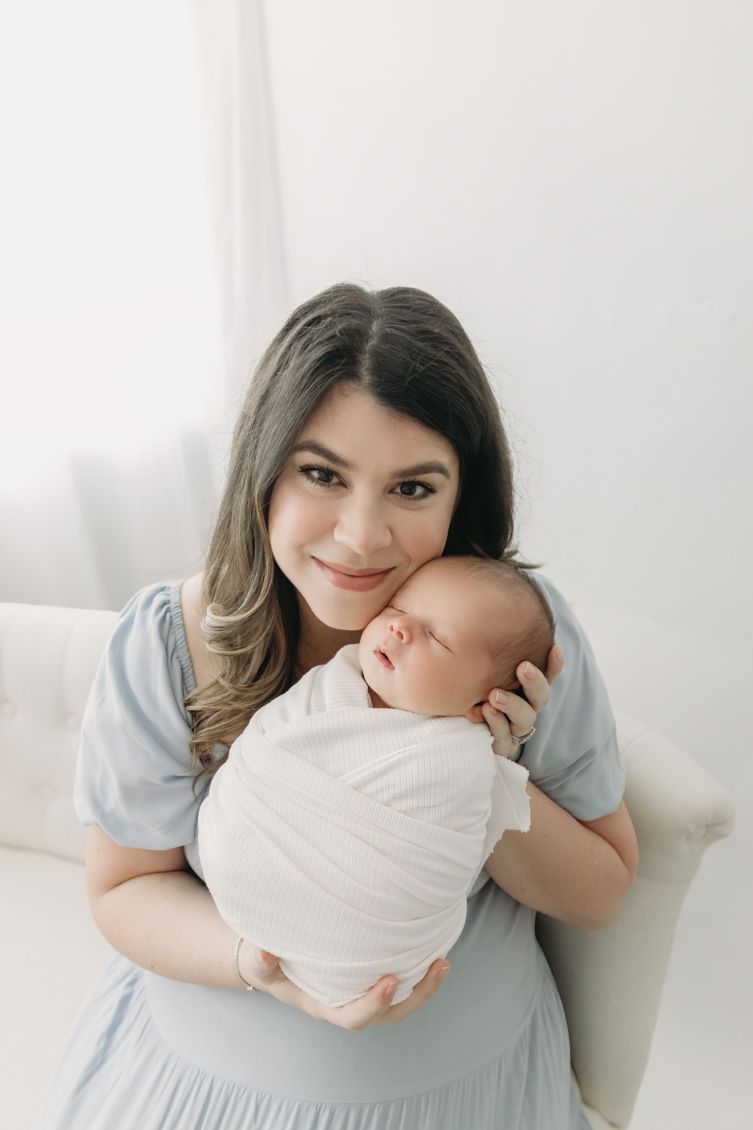 A tender moment as a young woman in a blue dress cradles a sleeping newborn baby swaddled in white.