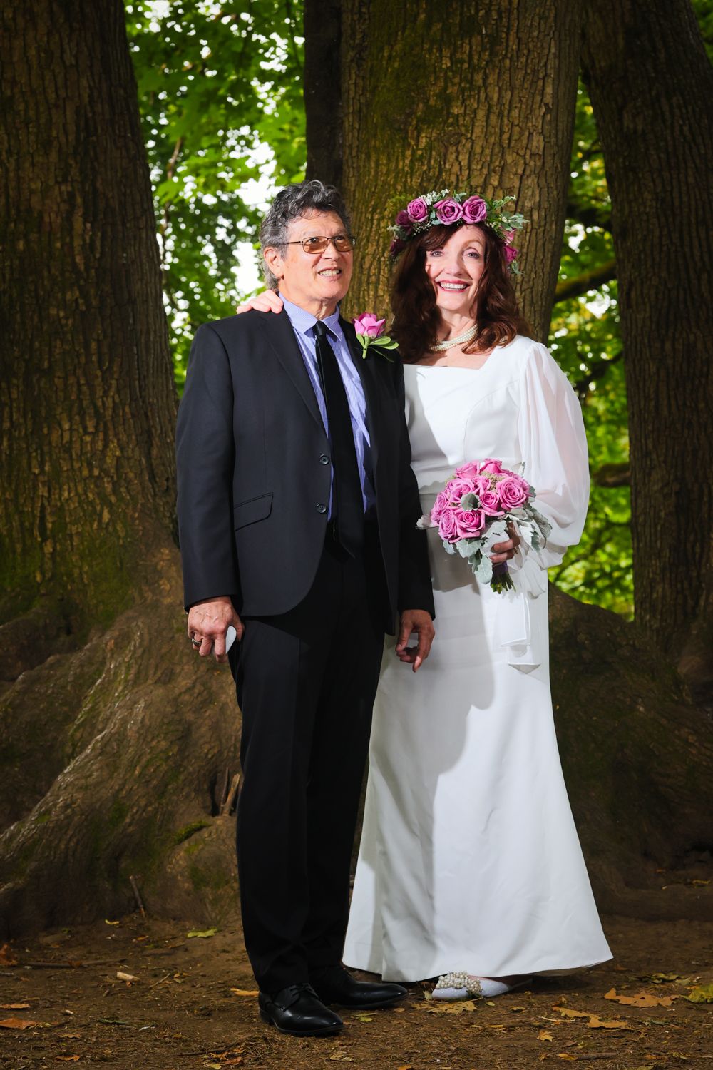 A couple stands together against a large tree trunk in an outdoor wedding setting with pink floral bouquet details.