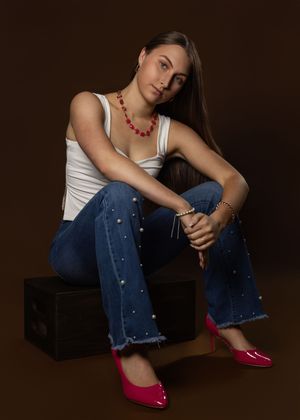 Model in white tank top, distressed jeans with pearl details, red flats and coral necklace poses on dark box against brown backdrop.