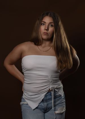 A person in a white ruched strapless top and distressed jeans poses against a dark background.