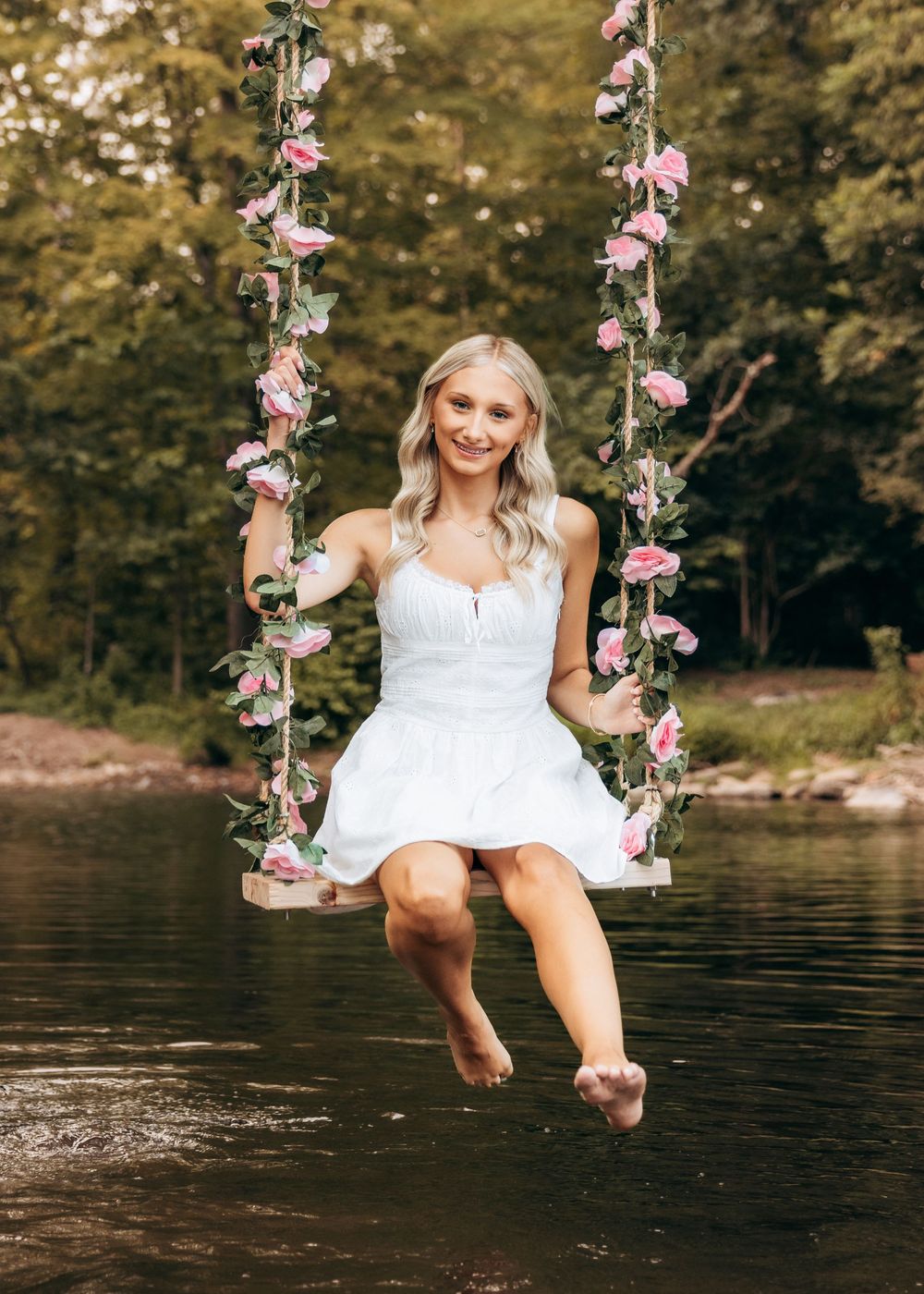 Whimsical photo series on a floral-decorated swing over water in white dress at sunset.