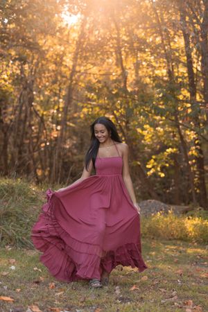 Woman in flowing burgundy dress twirls gracefully in autumn forest setting with golden sunlight filtering through trees.