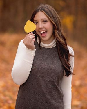 A woman in brown and white sweater holds up a golden autumn leaf while standing in a fall forest setting.