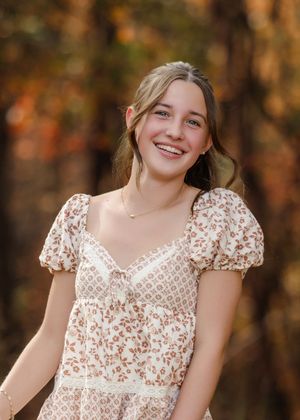 A person in a floral print dress poses outdoors during autumn with warm fall colors in the background.