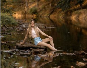 A person poses in denim shorts near a tranquil stream during golden hour, creating a dreamy outdoor portrait setting.