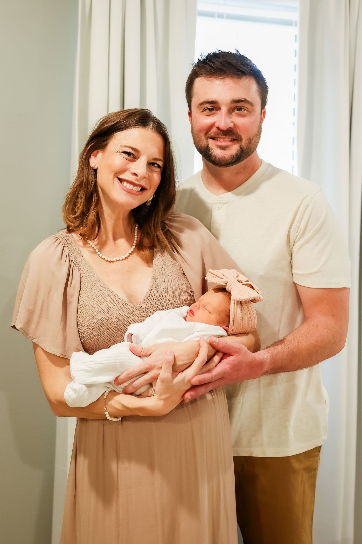A couple stands together indoors holding their newborn baby wrapped in white cloth.