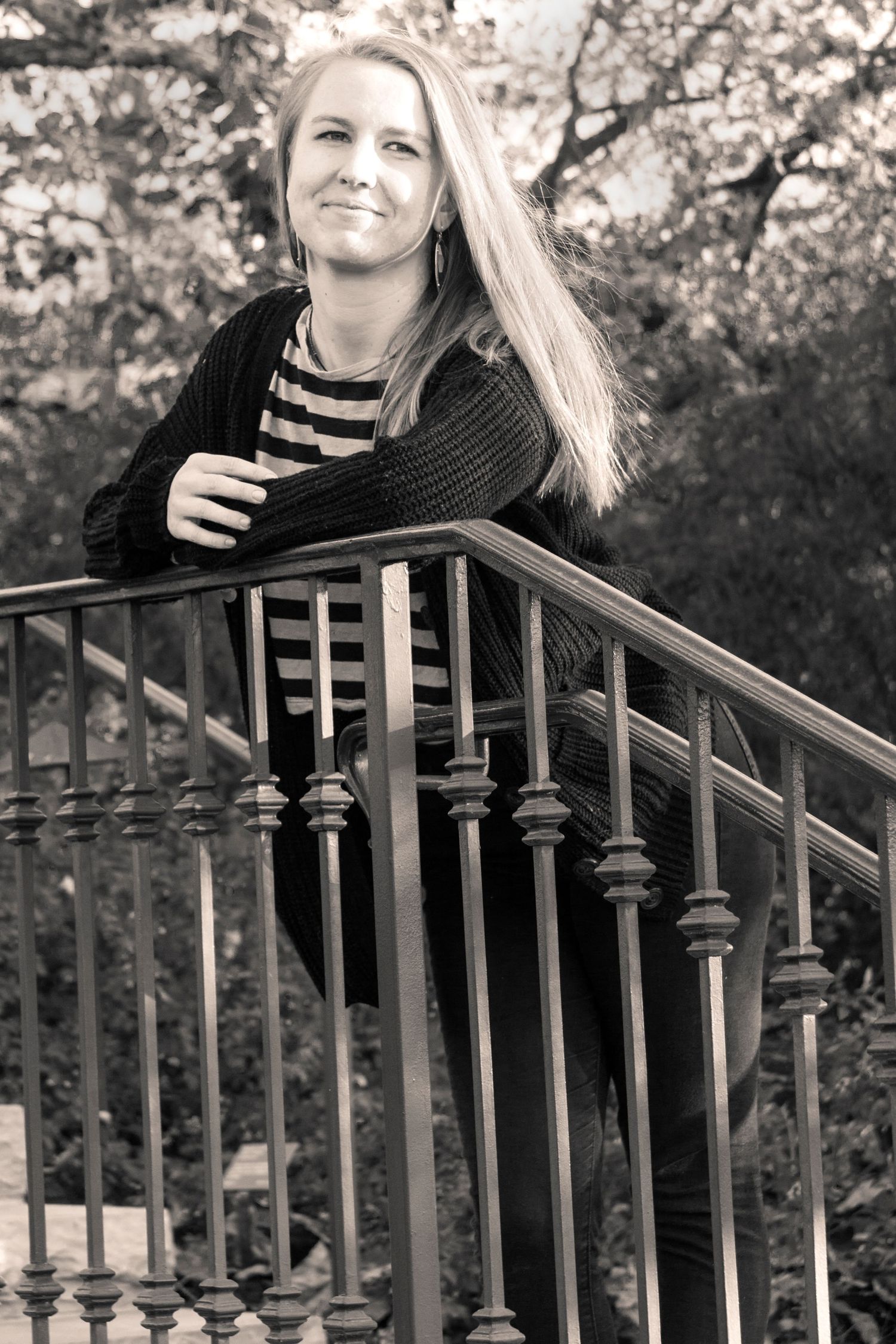 B&W Senior portrait of high school student with long blonde hair in stripped shirt and black sweater leaning on iron railing.