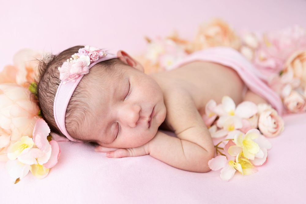A sleeping newborn baby lies peacefully among soft pink cherry blossoms and petals on a light pink background.