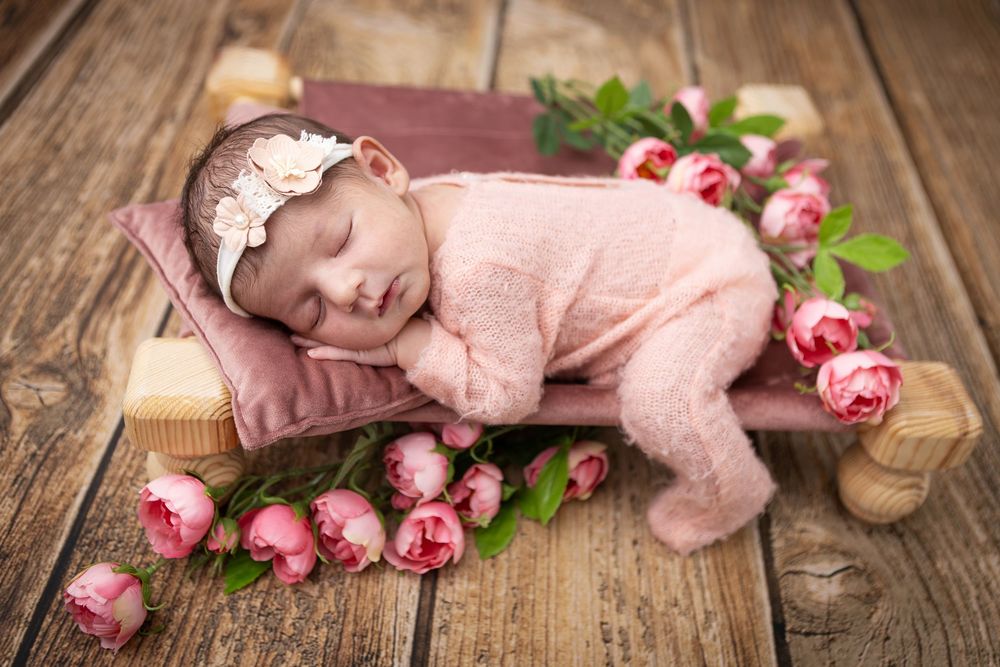 A sleeping baby in pink outfit lies on wooden surface surrounded by pink roses and wooden decorative elements.