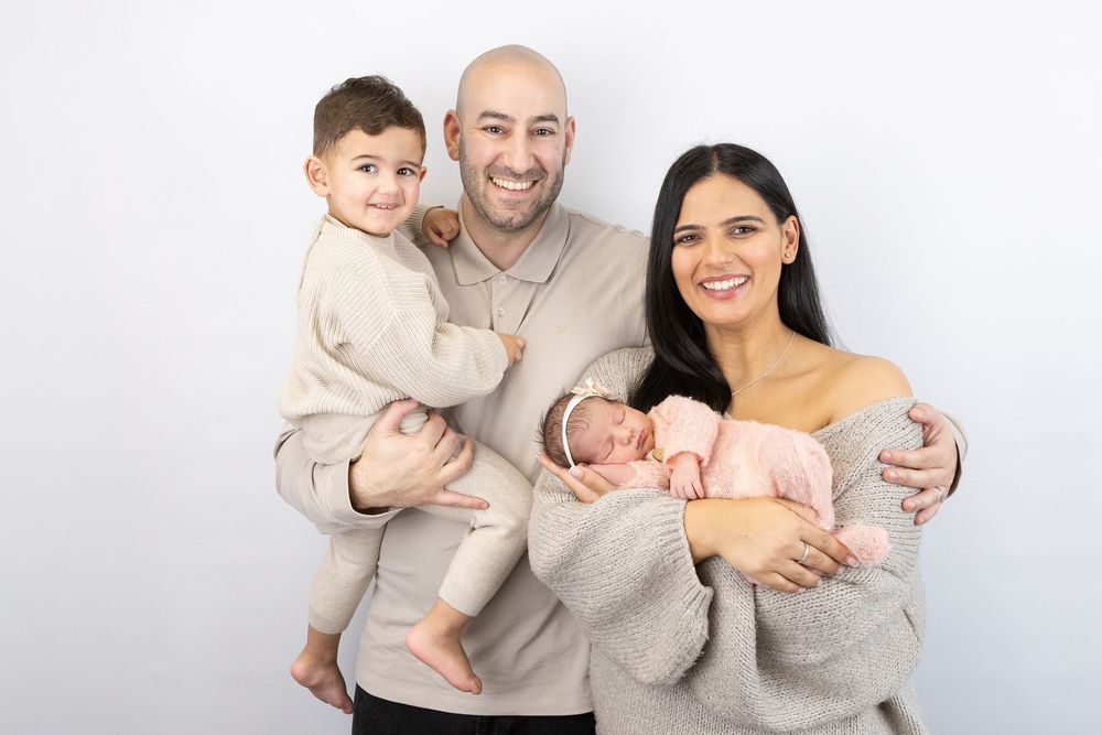 A happy family wearing neutral colored sweaters poses together with their newborn baby against a white background.