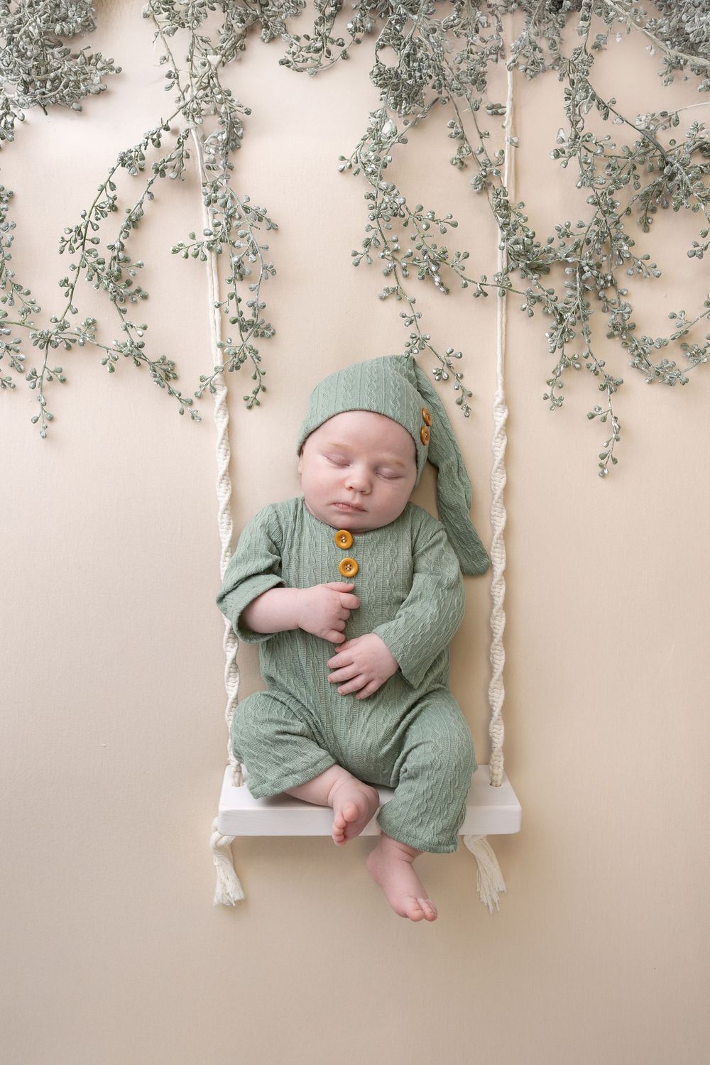 Baby in mint green outfit sleeping on white swing with delicate floral decorations above.