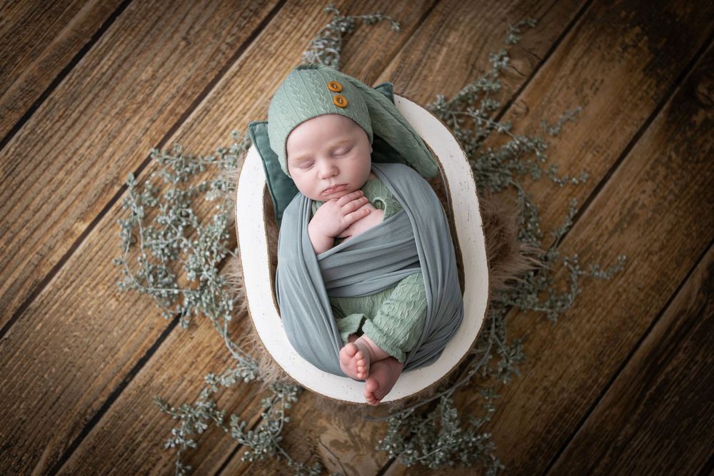 Sleeping newborn in sage green wrap nestled in white bowl with eucalyptus decor.