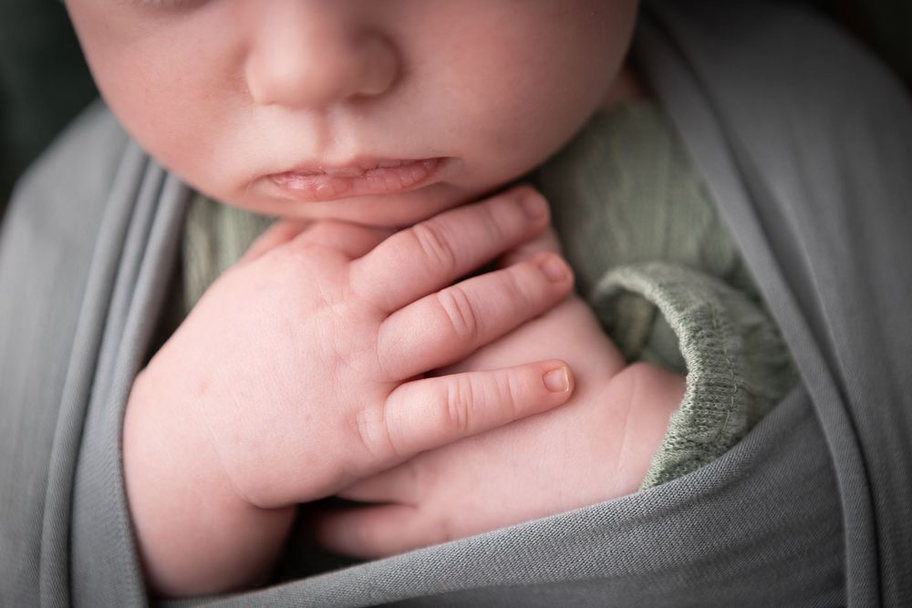 Newborn baby wrapped in soft gray fabric with delicate hands visible.