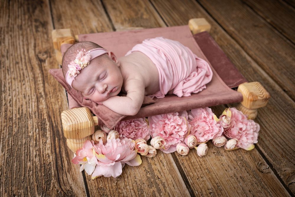 Sleeping newborn in pink wrap on wooden floor with floral decorations.