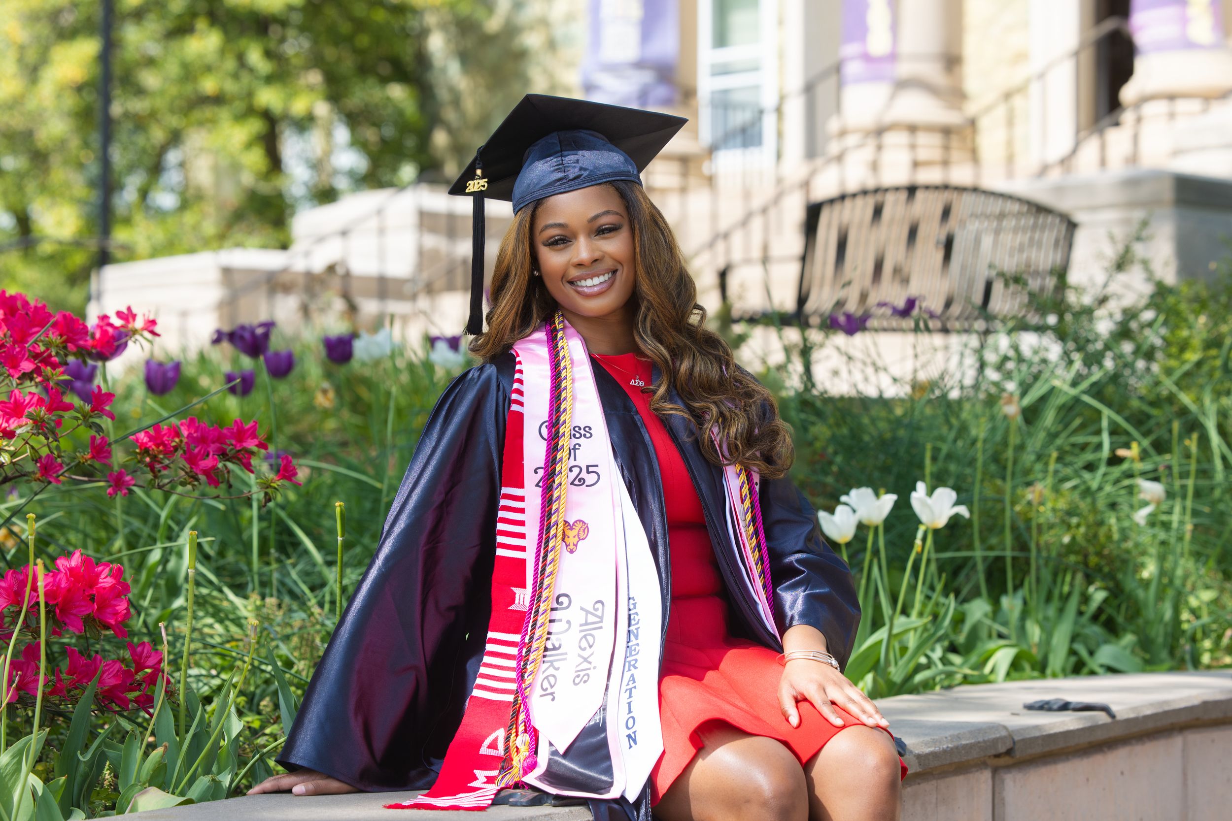 Graduate in cap and gown sitting on stone wall surrounded by blooming spring flowers in garden setting.