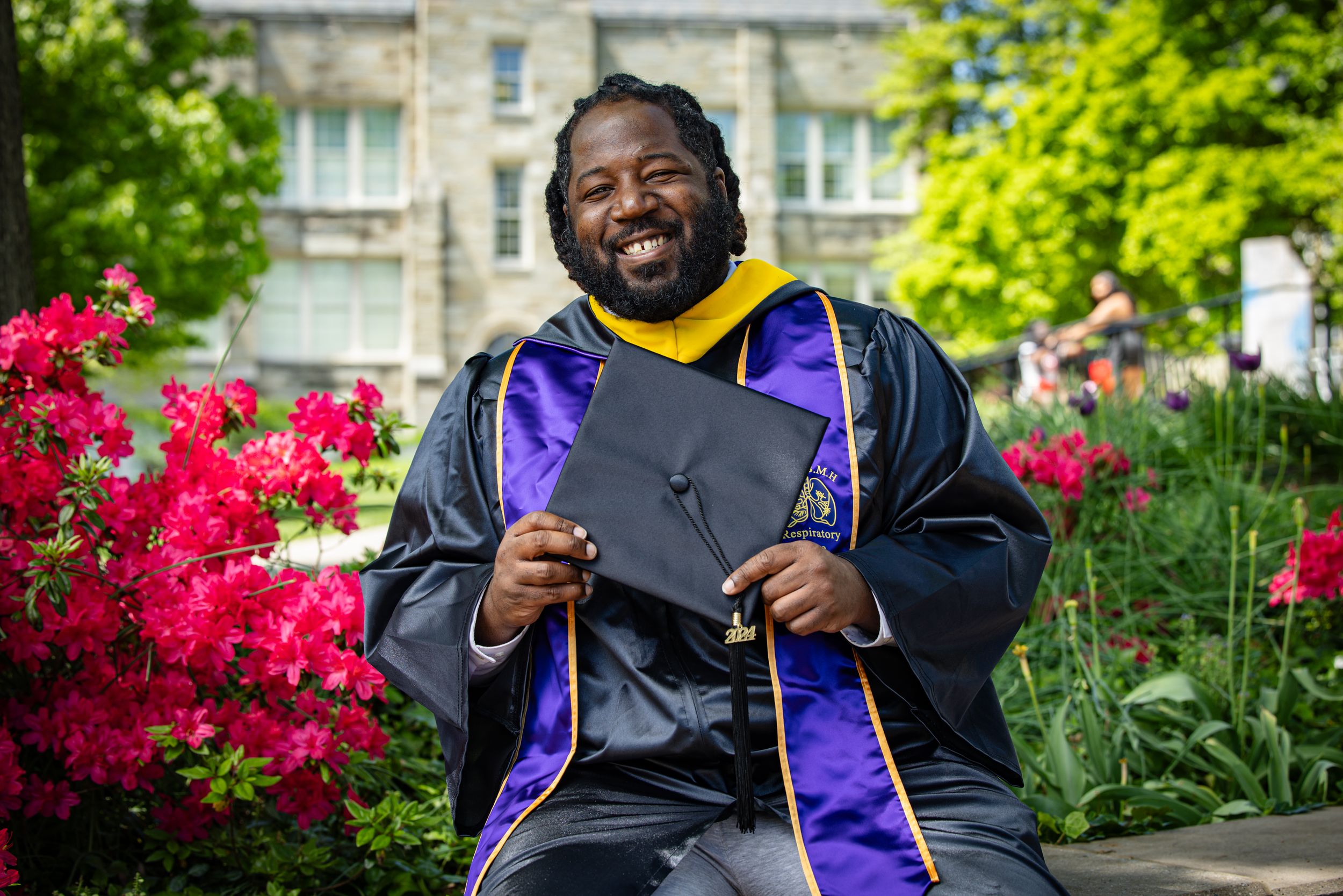 A joyful graduate in purple and black regalia holds up a diploma while sitting among blooming red azaleas.