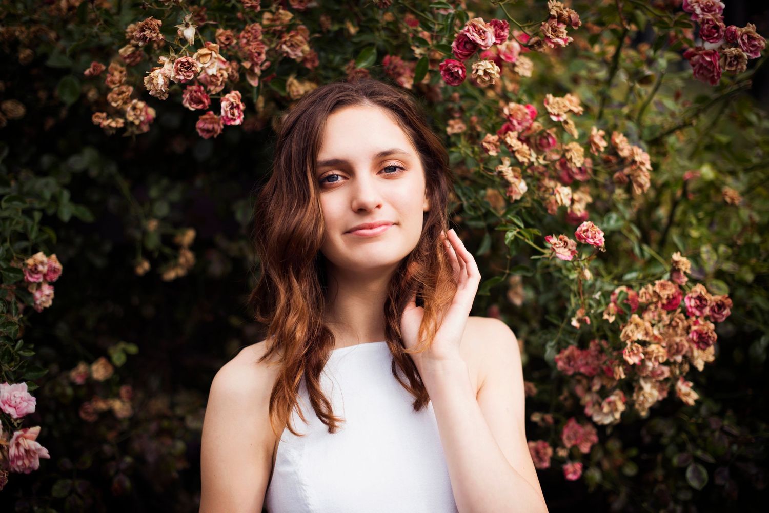 Young woman with wavy brown hair in a white top, standing amid blooming rose bushes, gently touching her hair at the Berkeley Rose Garden.