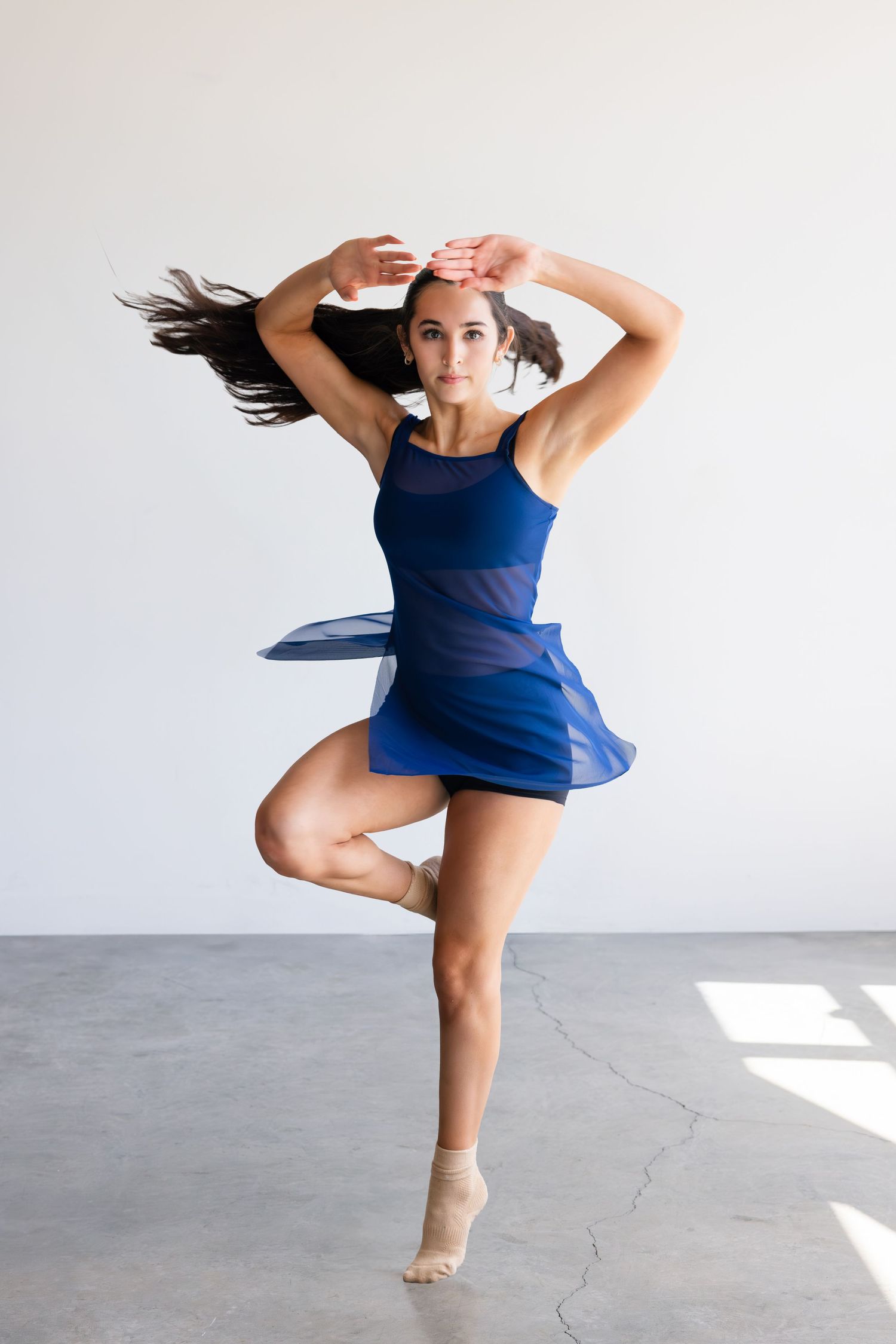 Dancer in blue leotard dress performs dynamic ballet pose with flowing movement on a concrete studio floor.