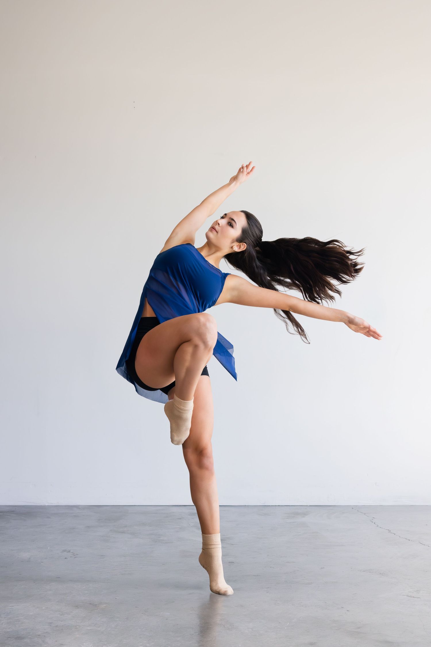 Contemporary dancer in flowing blue dress performs graceful leap with long dark hair in motion against light background.