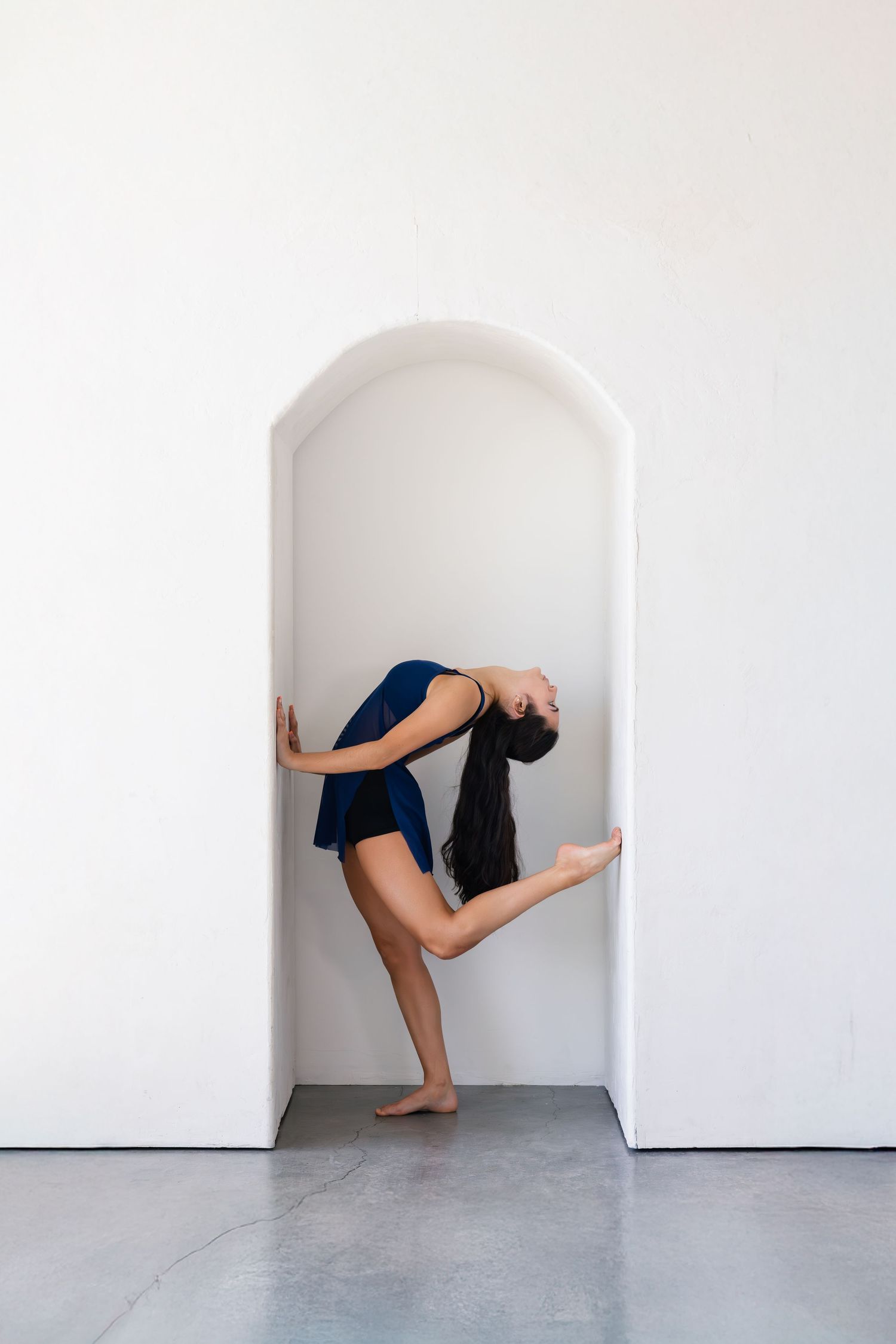 A graceful dance pose showing flexibility as performer arches backwards in a white alcove wearing blue.