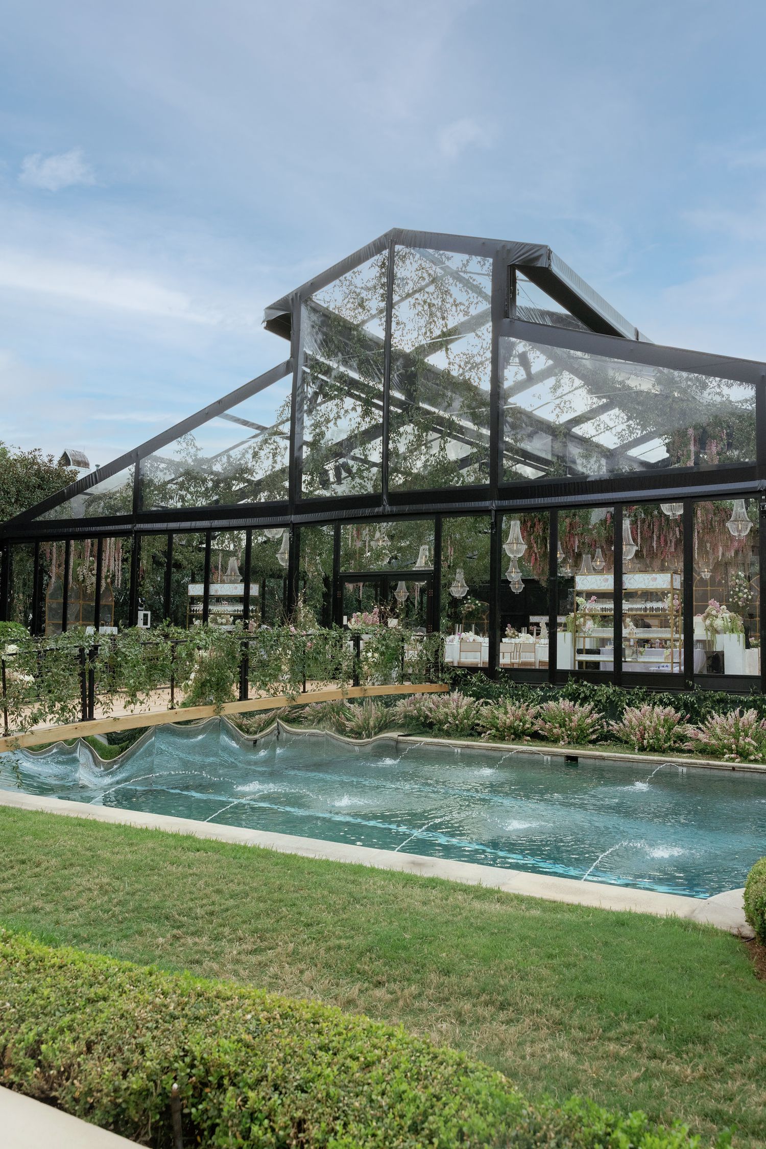 A modern glass greenhouse structure with a sleek swimming pool in front surrounded by landscaped gardens and grass.