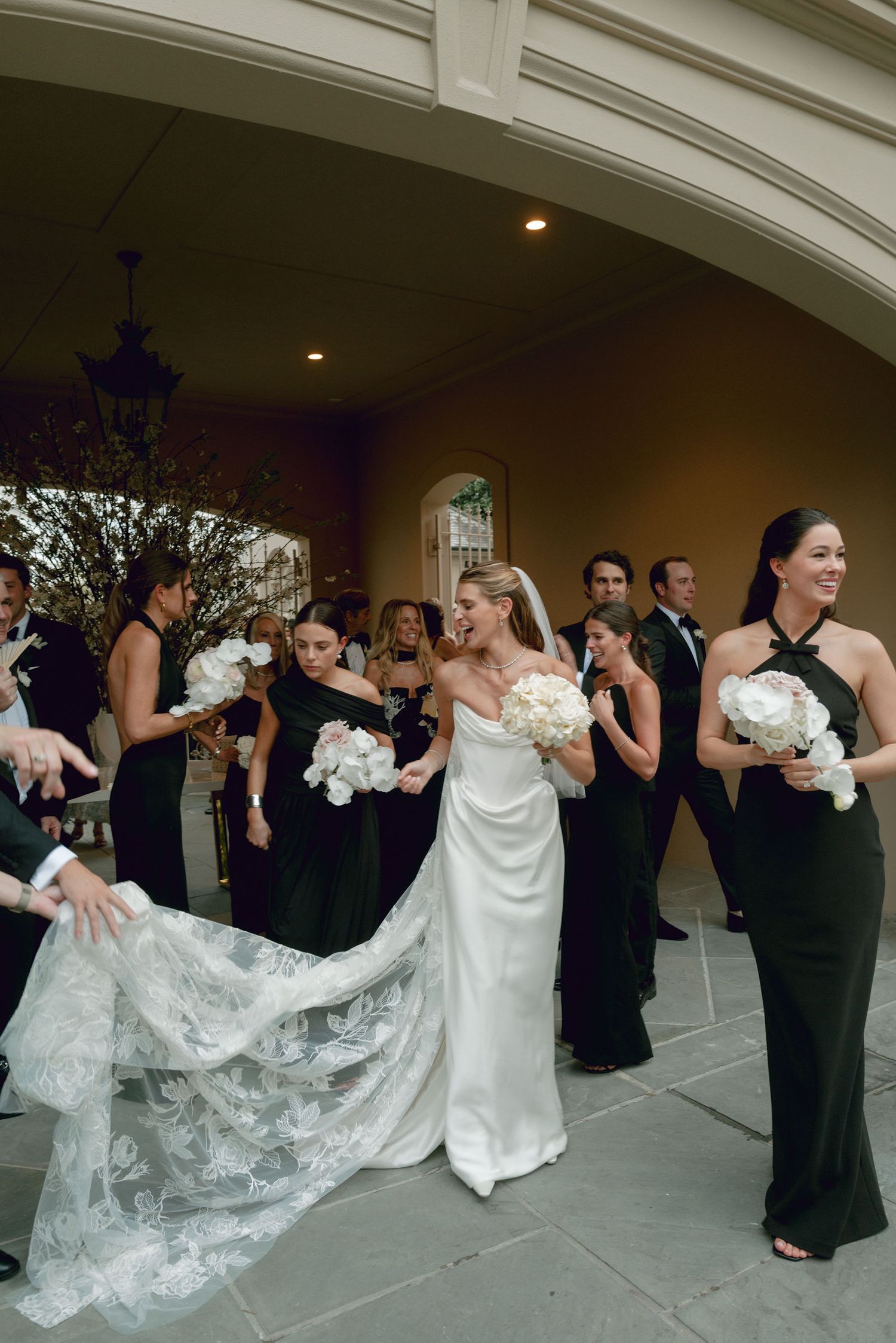 Bridal party in elegant black dresses and a white wedding gown with lace train gathered outside an arched doorway.