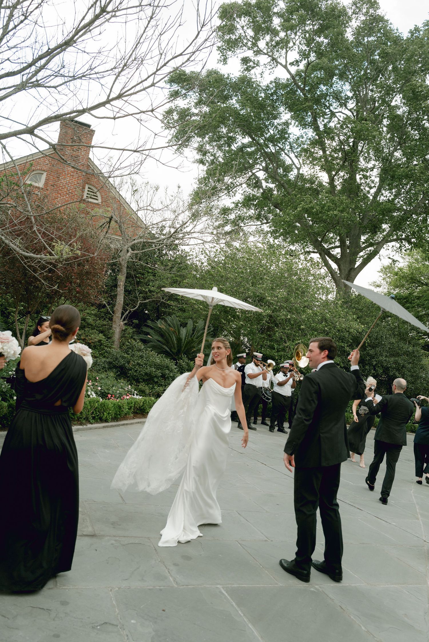 Wedding celebration taking place on a stone patio with green trees and brick building in the background.