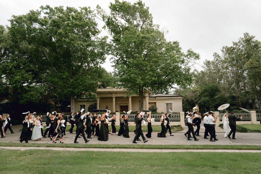 A line of formally dressed wedding guests with umbrellas walk in procession along a garden path during an outdoor event.
