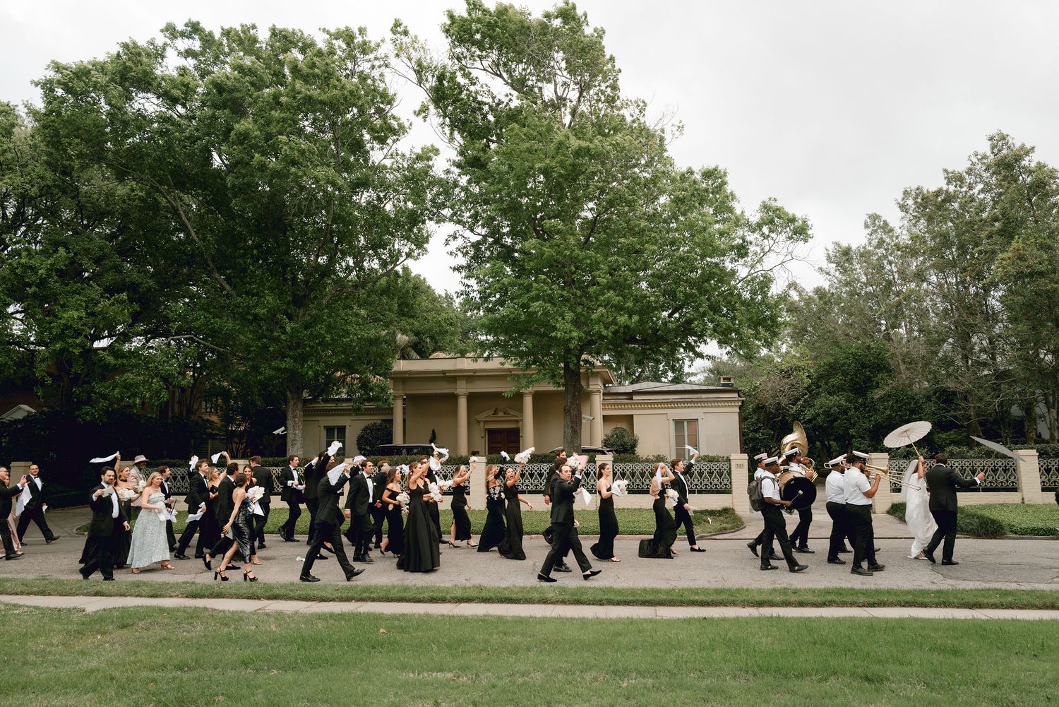 A line of formally dressed wedding guests with umbrellas walk in procession along a garden path during an outdoor event.