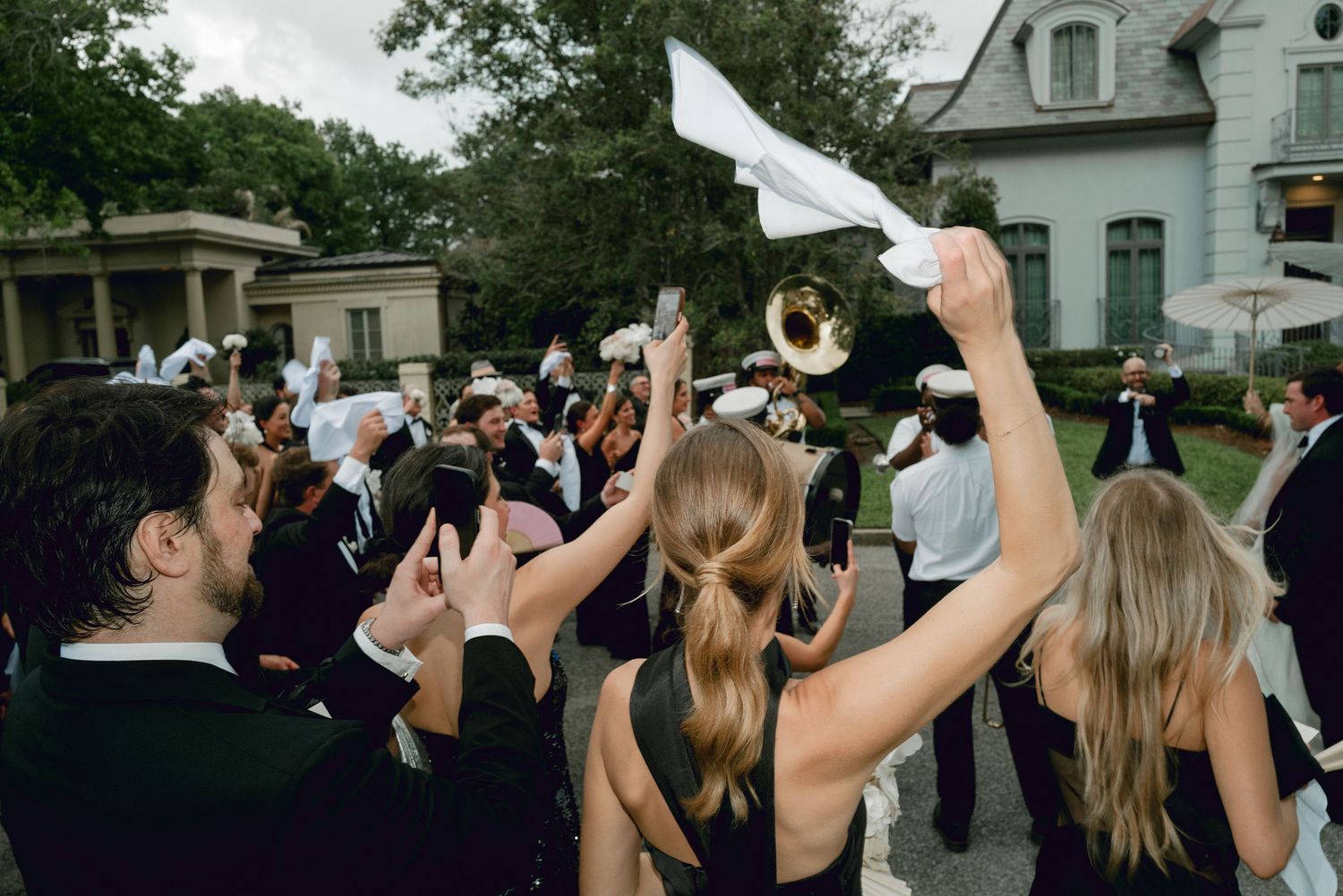 A crowd of formally dressed people wave white napkins and cheer during an outdoor celebration in front of a historic building.