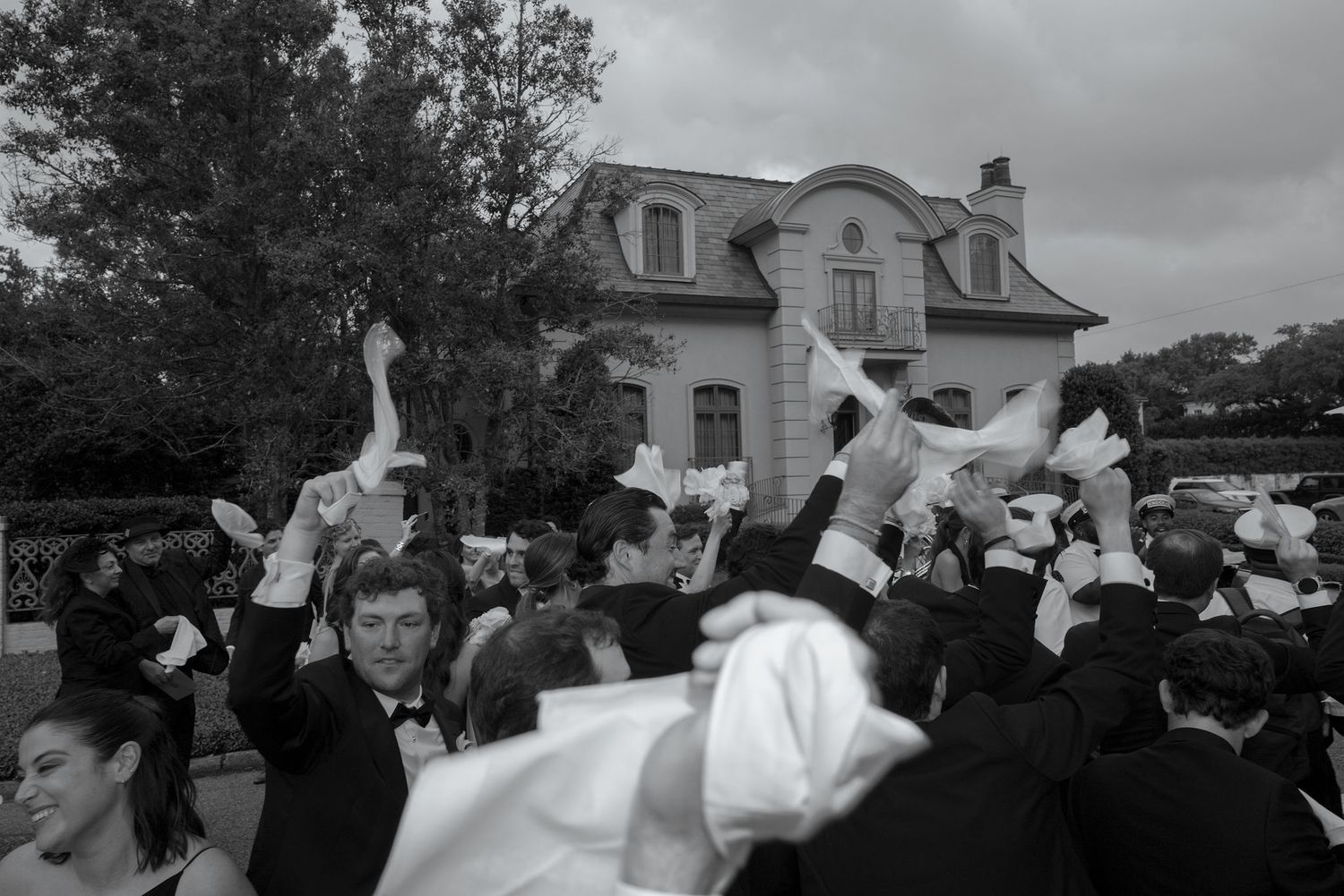 A celebratory crowd throws their hands up in jubilation outside a mansion with dormer windows.