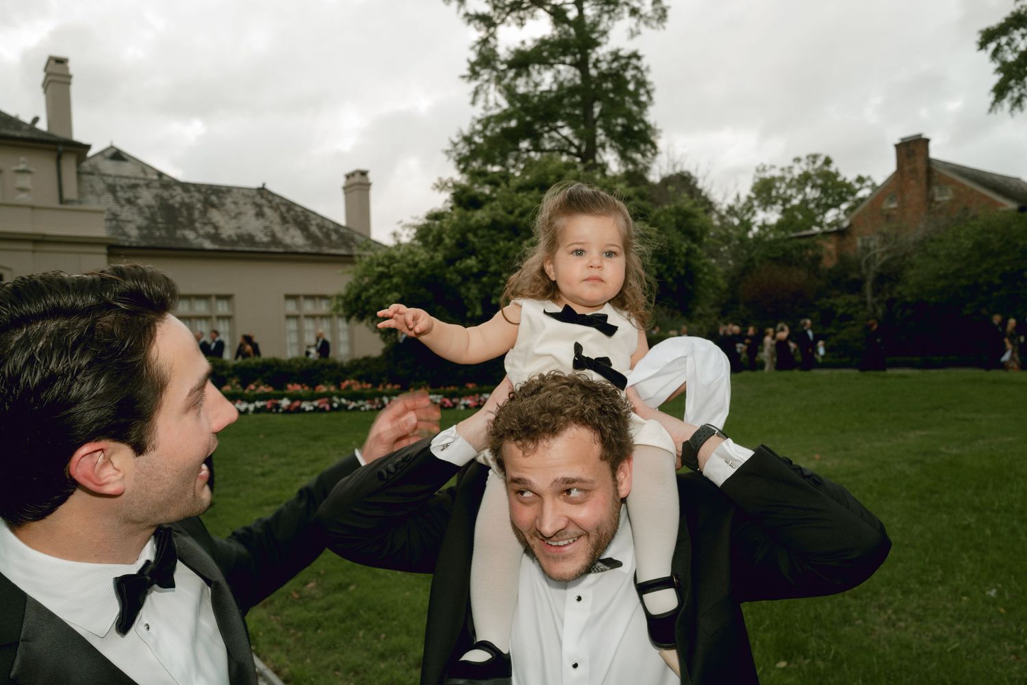 Two people in tuxedos at an outdoor celebration with someone sitting on their shoulders in a garden setting.