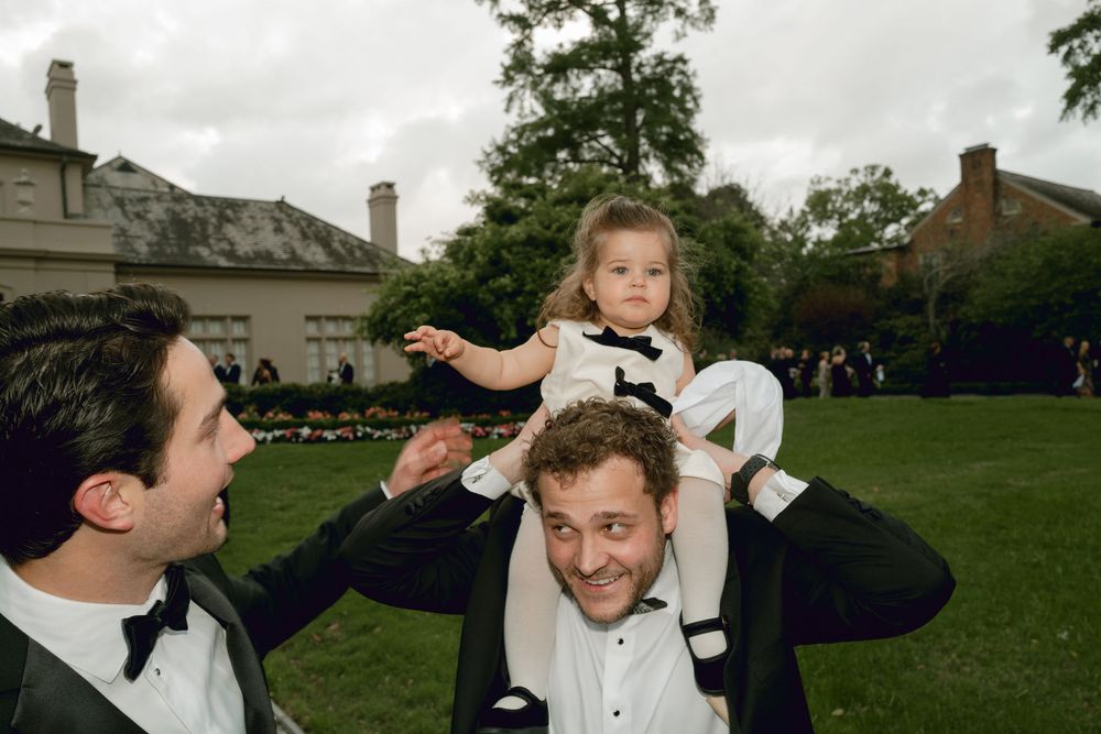 Two people in tuxedos at an outdoor celebration with someone sitting on their shoulders in a garden setting.