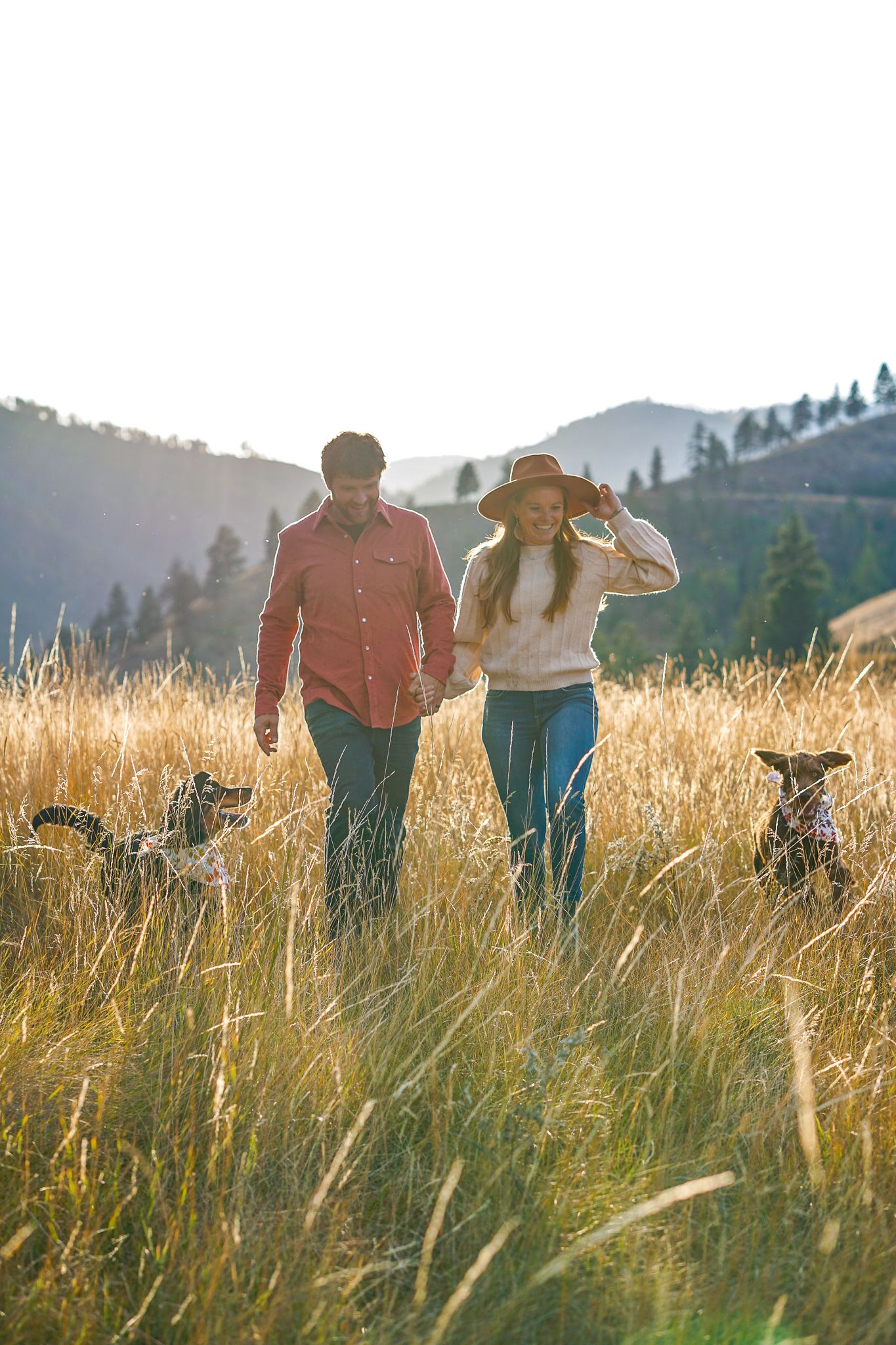 Two people walk hand in hand through golden grass on a hillside at sunset, silhouetted against a misty mountain landscape.
