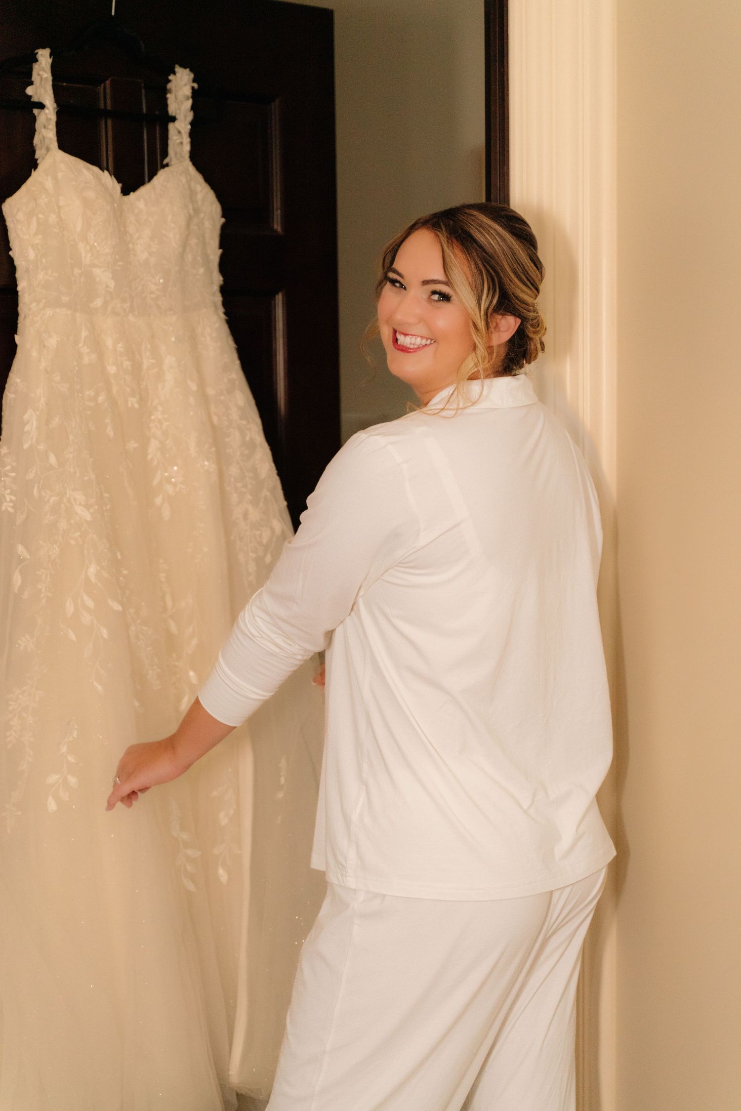 A person wearing a white robe admires a detailed lace wedding gown displayed against a dark door.
