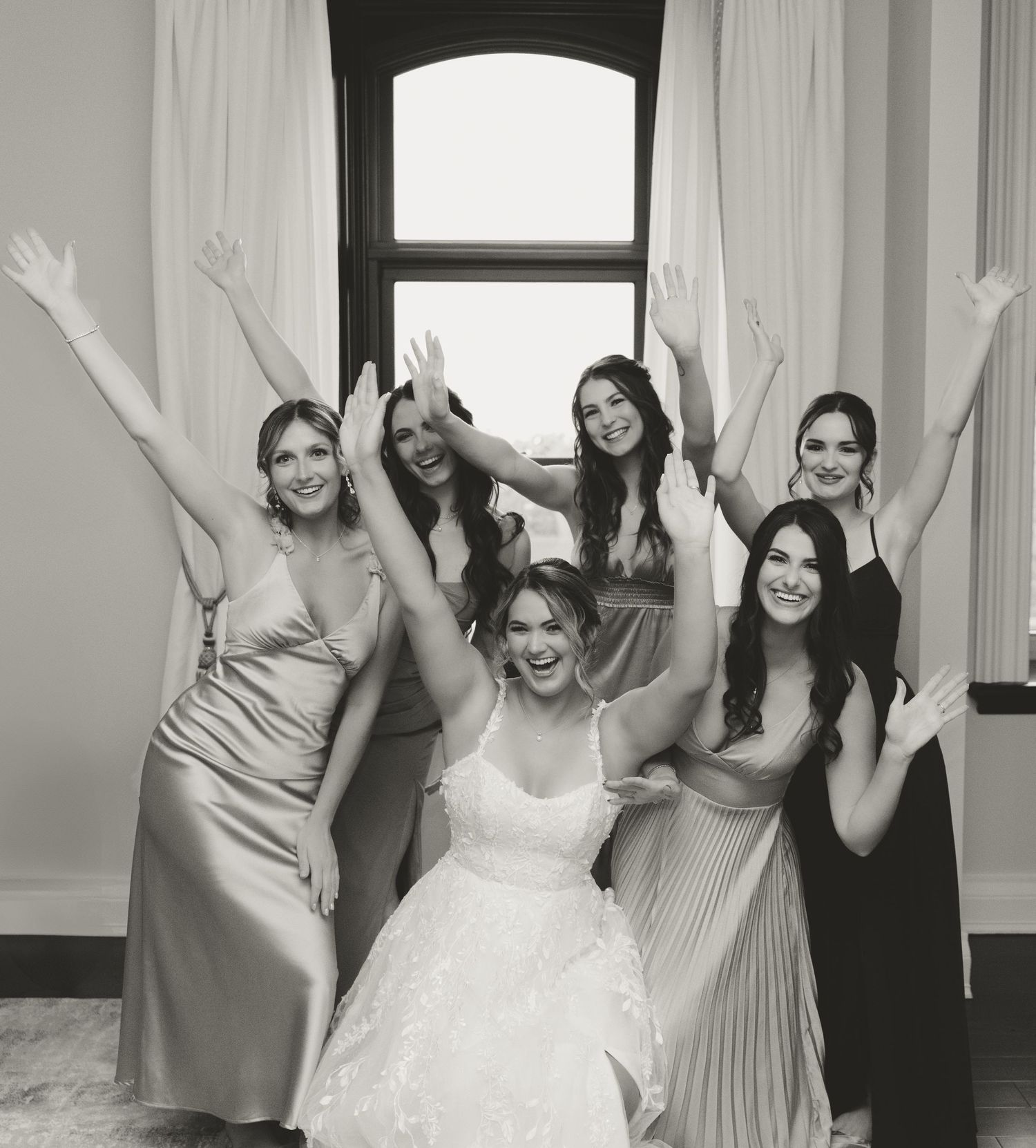 Black and white photo of bridal party with raised arms in elegant dresses by a window.