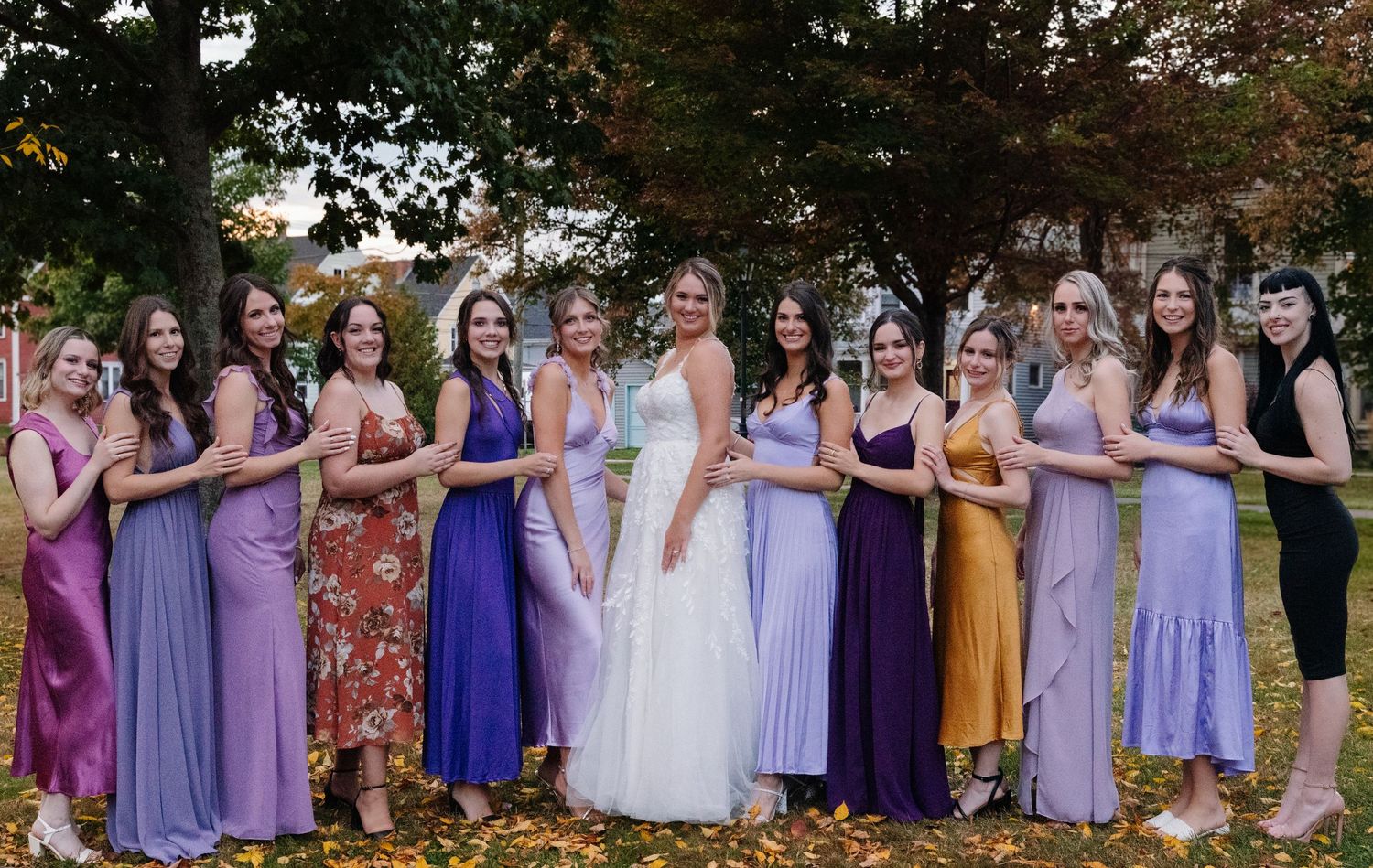 Bridal party in varying shades of purple dresses poses together against fall foliage.