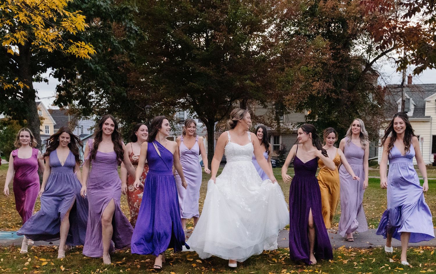 Bridal party wearing various shades of purple dresses posing on fall lawn.