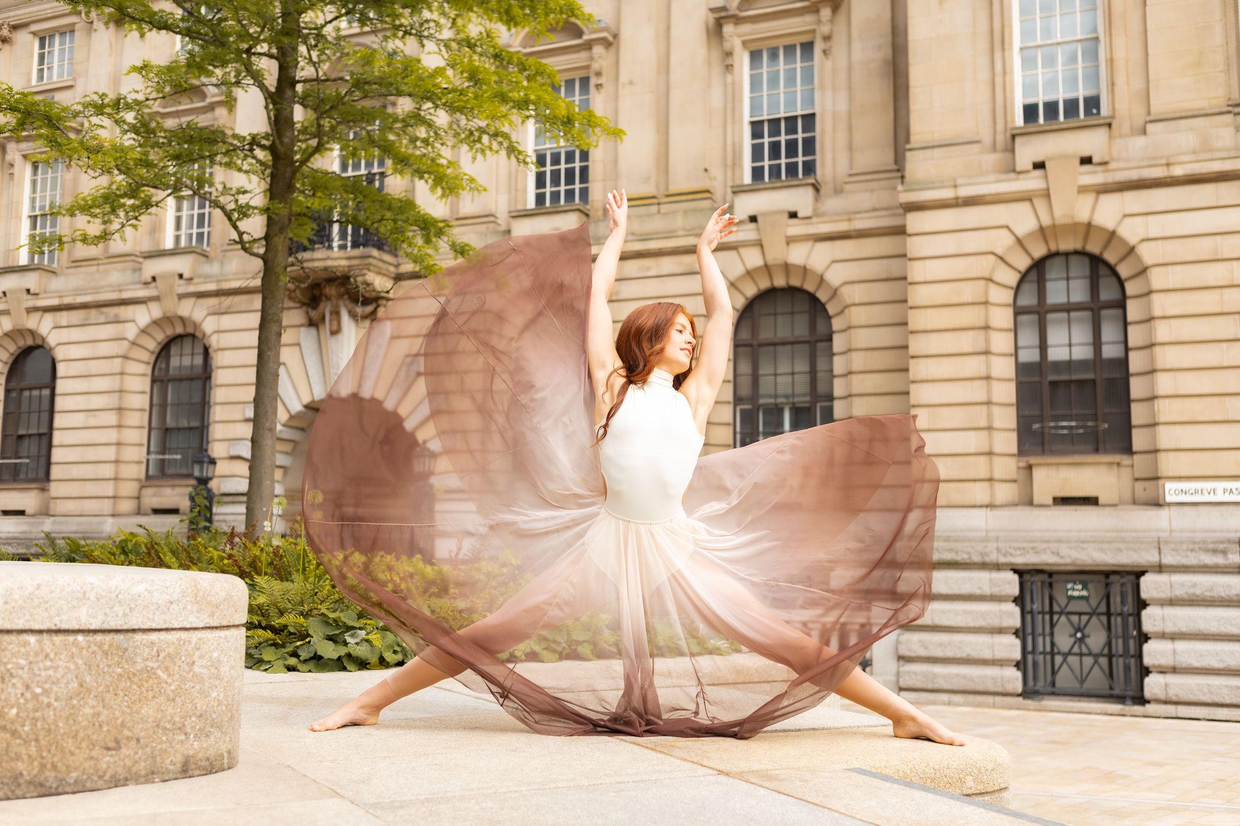 A graceful dancer in a flowy dress performs a dynamic movement against the backdrop of a classical stone building.