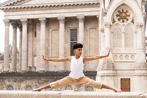 A ballet dancer performs a graceful split leap in front of an ornate historical building with classical architecture.