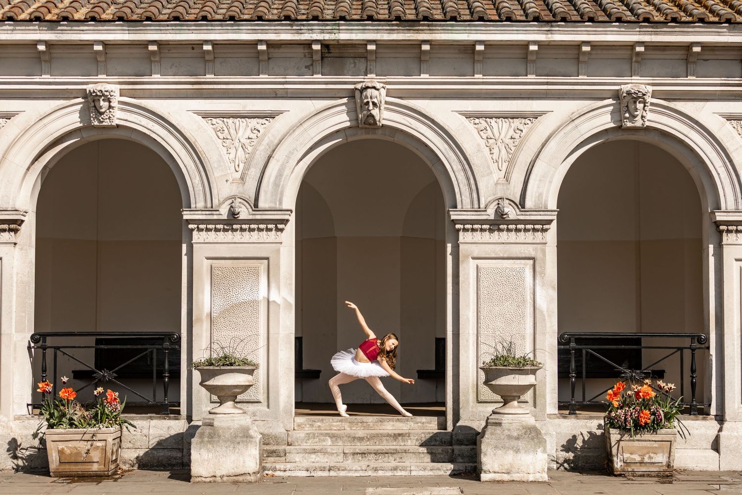 Ballet dancer performs graceful leap pose between ornate classical stone archways with potted flowers.