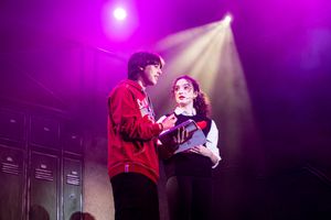 Two performers sing together on stage under dramatic purple lighting during a theatrical production.