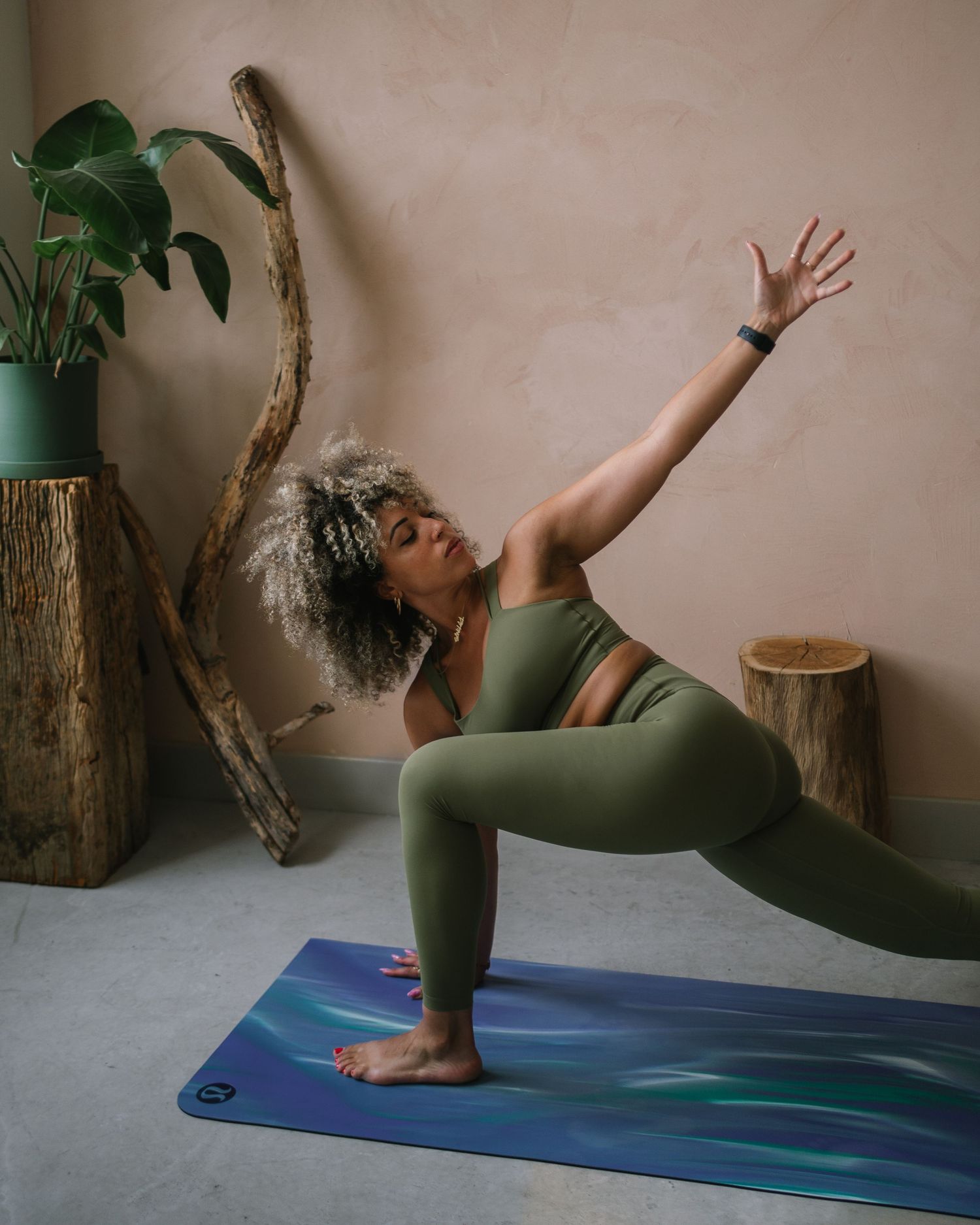 A person in olive green athletic wear performs a low lunge yoga pose on a blue mat next to potted plants and wooden decor.