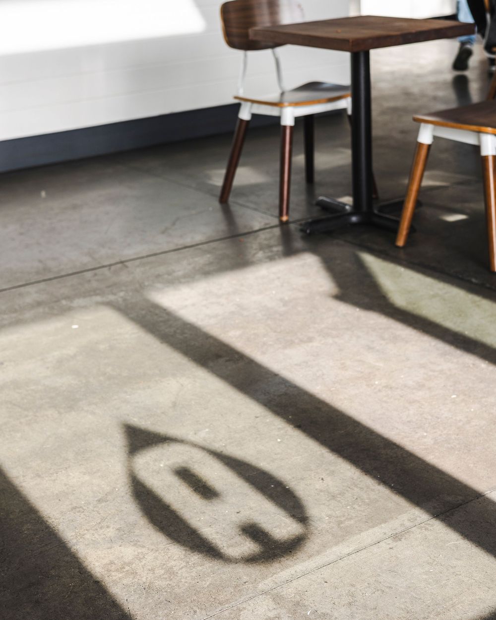 Shadow cast on concrete floor shows 'A' letter typography next to cafe table and stools.